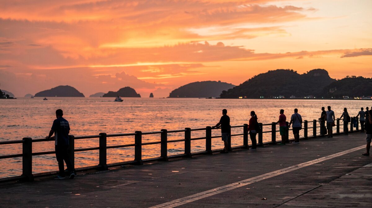 The Kota Kinabalu waterfront promenade at sunset, silhouetted figures along the rail, the sky burning orange over the South China Sea and the islands.